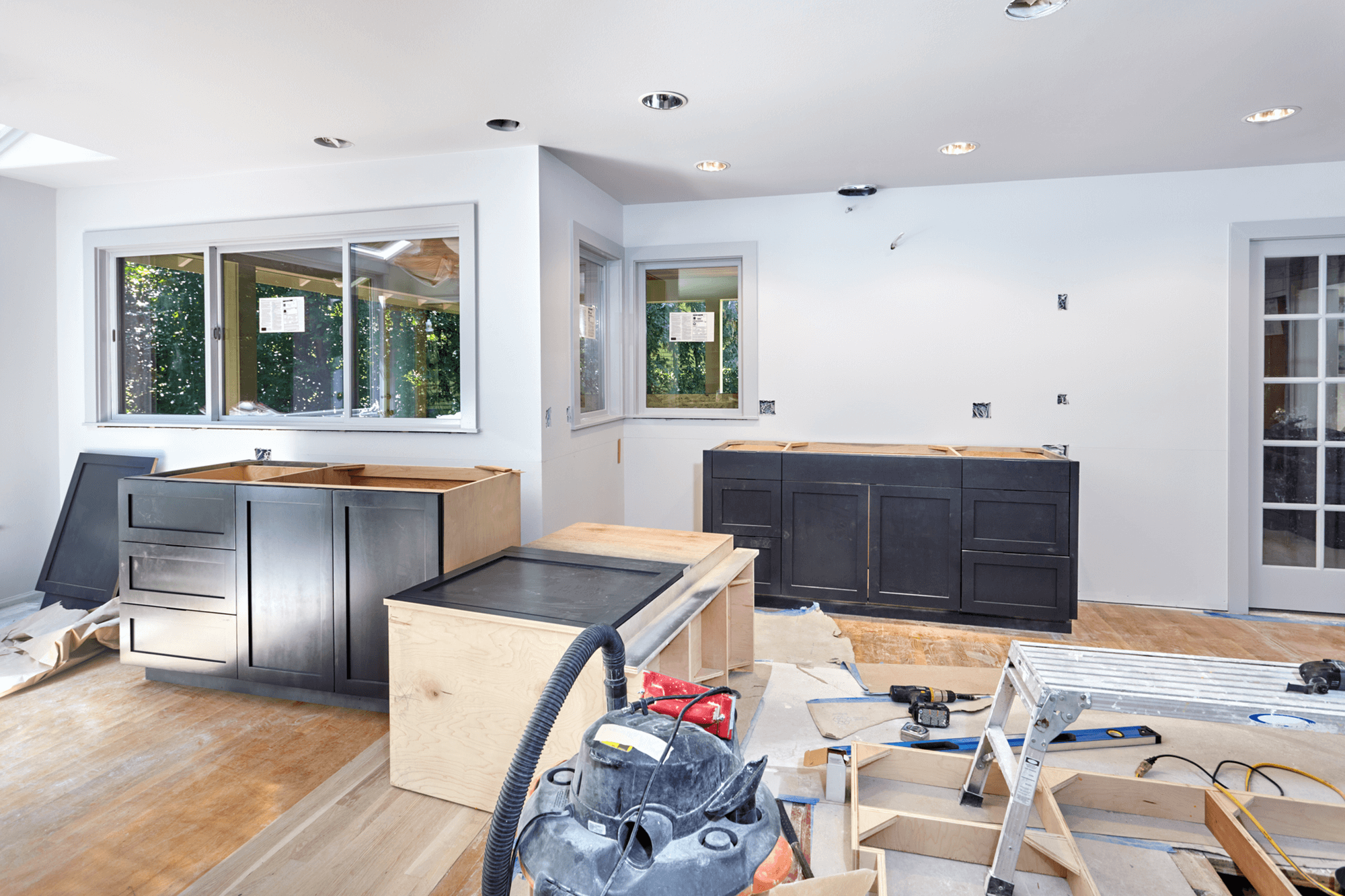 Modern home renovation in progress featuring dark cabinet boxes, light wood countertops, recessed ceiling lights, and large windows overlooking greenery